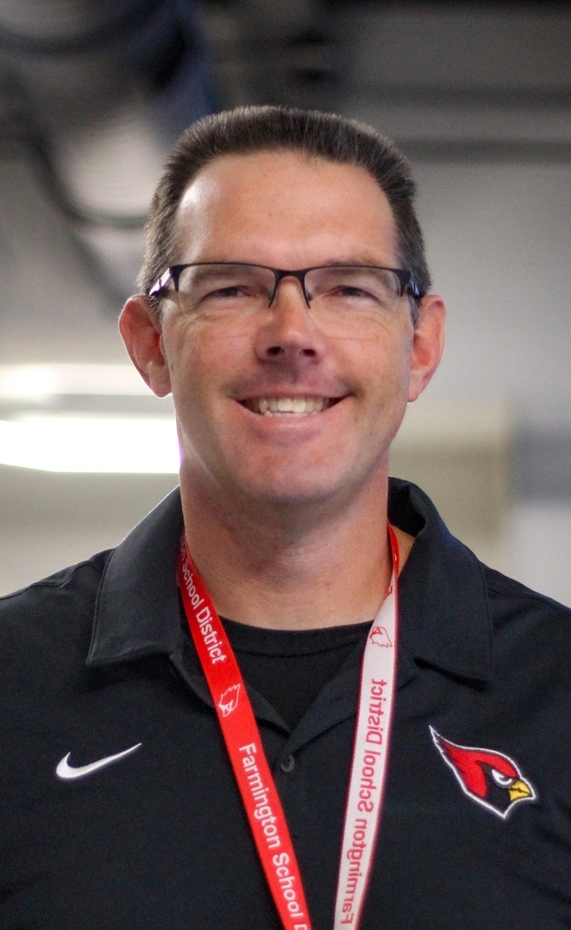 Man in glasses smiles in a room; wearing black shirt with red lanyard with text and a red bird.