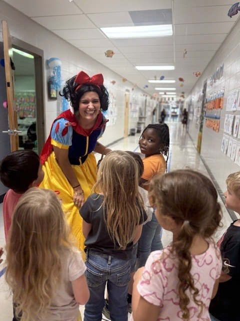 A woman dressed as Snow White talks to a group of children in a school hallway.