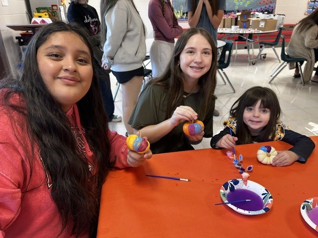 Three girls sitting at an orange table with crafts on it. They each hold painted pumpkins.