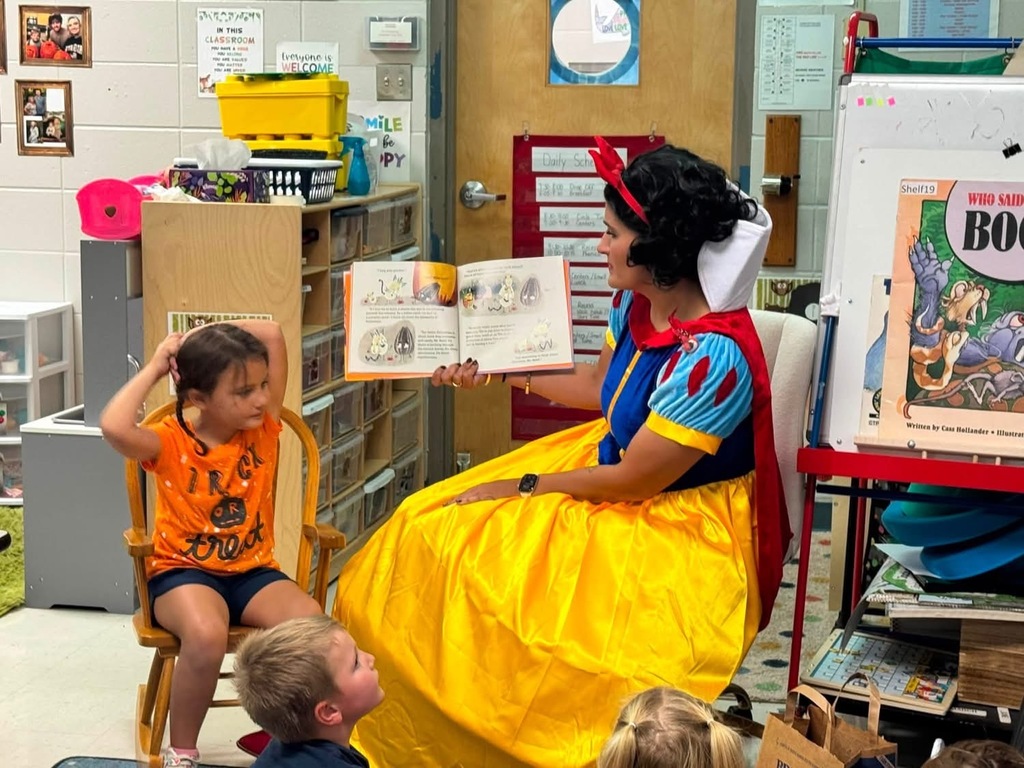 Woman dressed in Snow White costume reads story to children sitting in chairs; colorful books on shelves.
