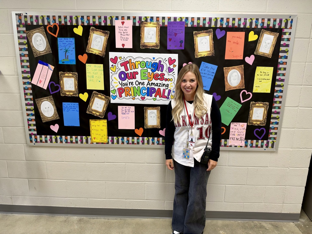 Person stands in front of bulletin board with colorful papers and pictures, including a principal photo.