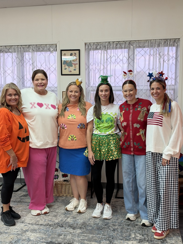 Adult females in festive costumes stand together in a room with windows and a framed picture.