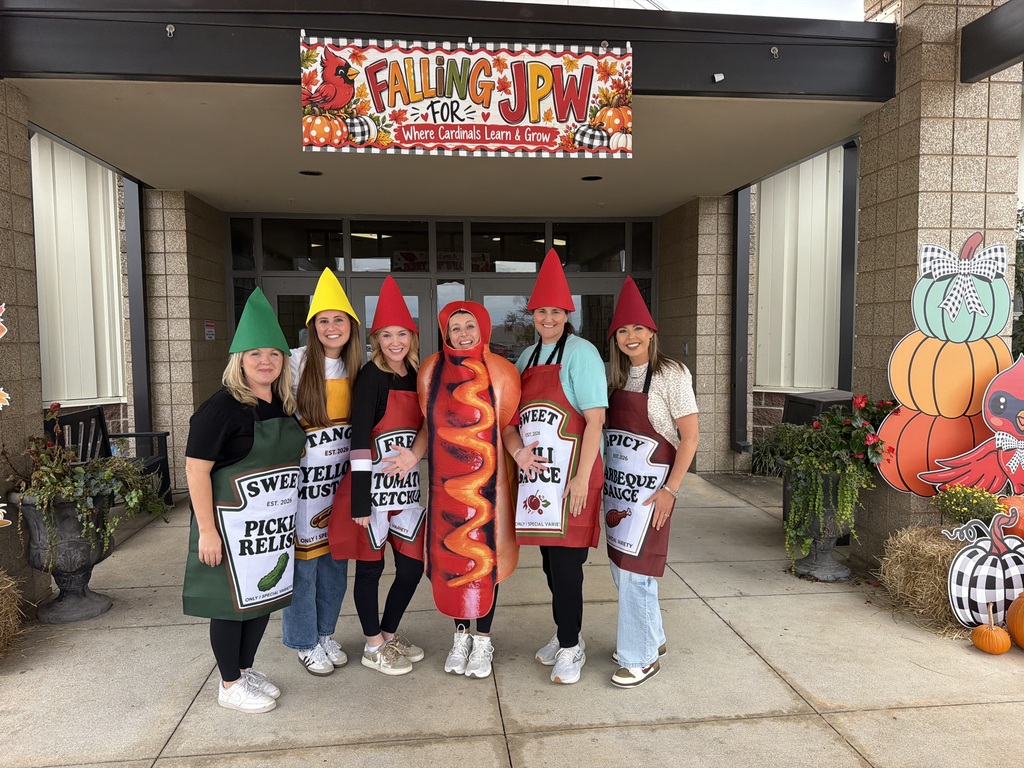 Group of students wearing costumes and aprons stand in front of a building with pumpkins and gourds.