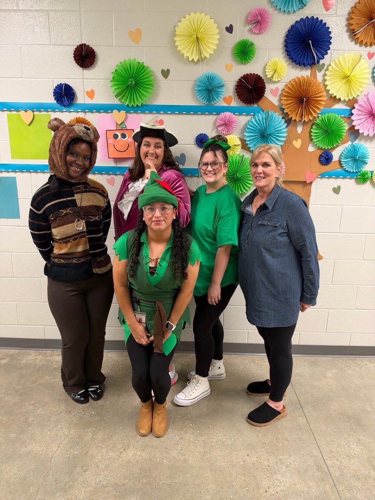 Five people in costumes pose for a picture in a classroom with a colorful paper tree and flowers on the wall.