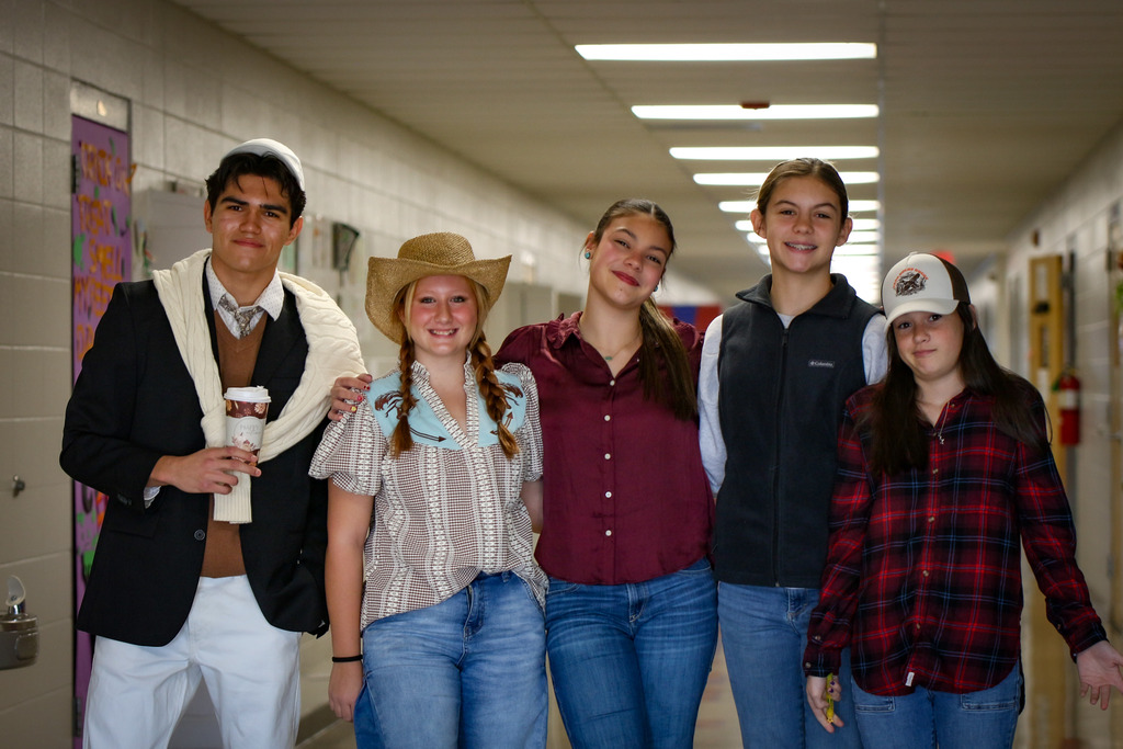 Five students dressed in various costumes stand in a hallway, smiling for a photo. One holds a drink, and another wears a cowboy hat.