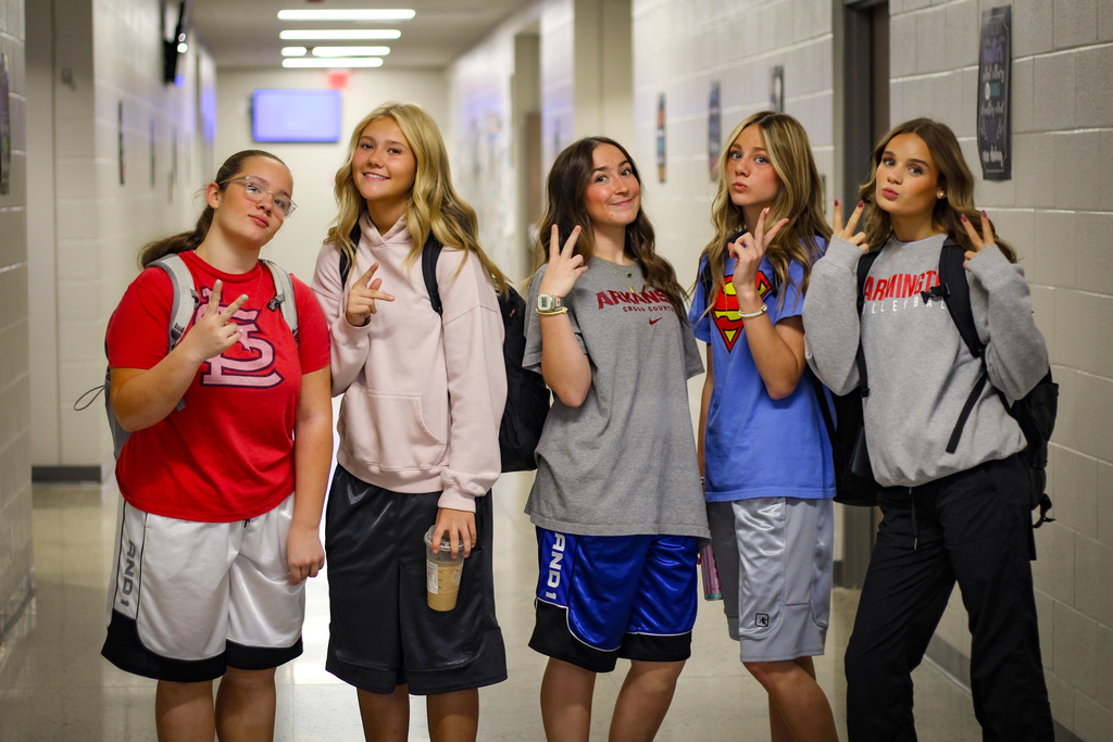 Five girls in school uniforms pose for a photo in a hallway, making peace signs with their hands.