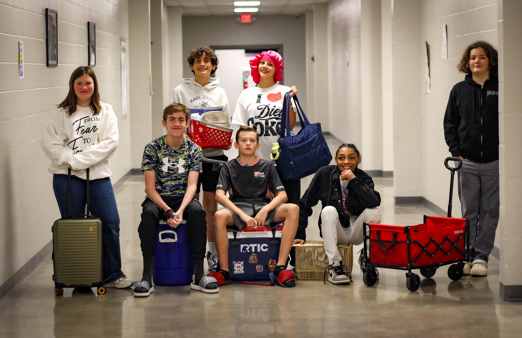 A group of students in a hallway, some seated on coolers, some standing, with luggage and a stroller.