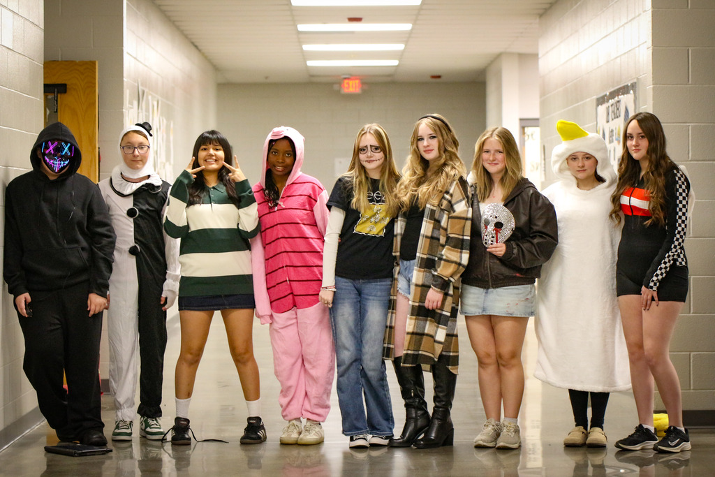 Students dressed in costumes stand in a hallway, looking forward. The hallway has white walls and gray floor.