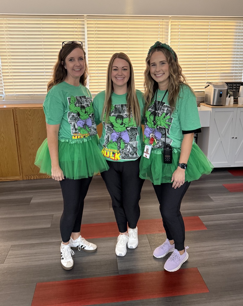 Three women in green T-shirts with superhero designs and tutus stand in a room with a wooden desk.