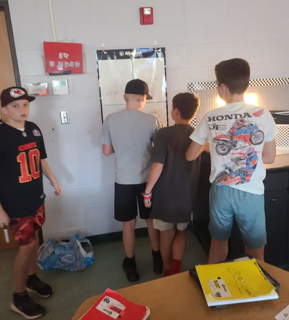 A group of students are gathered around a whiteboard, actively collaborating on an activity involving a large coordinate plane grid.