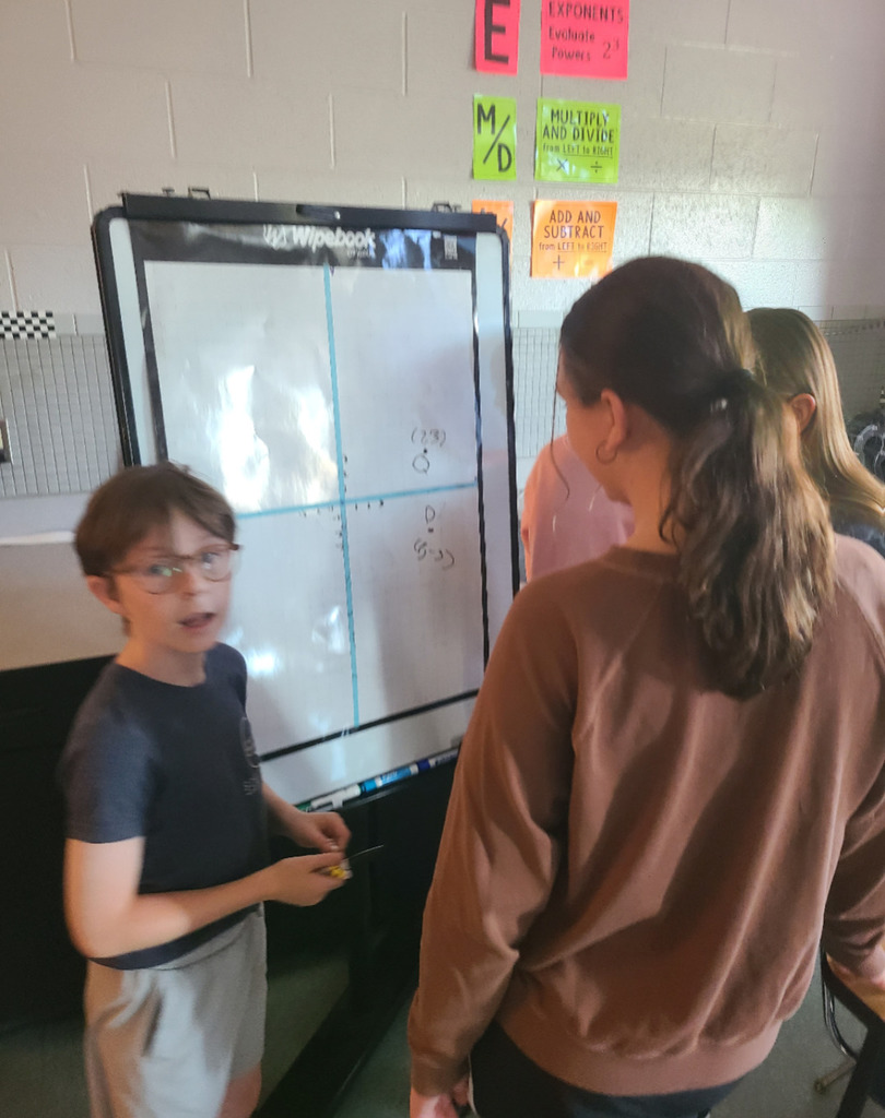 A group of students are gathered around a whiteboard, actively collaborating on an activity involving a large coordinate plane grid.