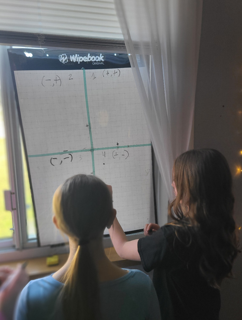 A group of students are gathered around a whiteboard, actively collaborating on an activity involving a large coordinate plane grid.