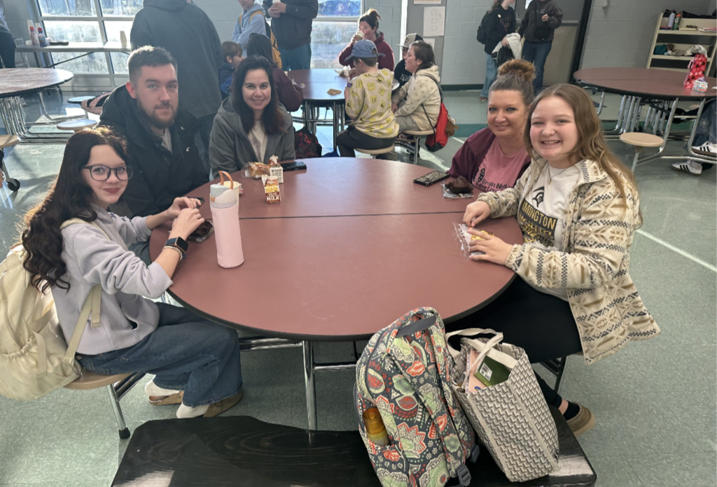 Students and their families sitting together at a table, smiling and enjoying muffins and coffee in the school cafeteria.