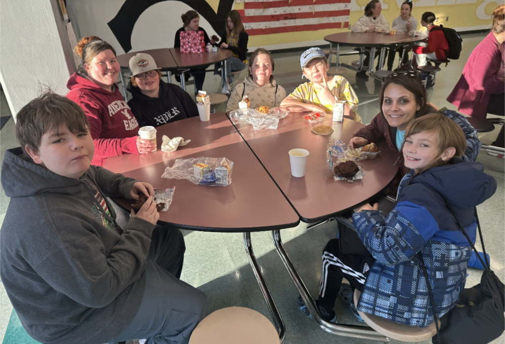 Students and their families sitting together at a table, smiling and enjoying muffins and coffee in the school cafeteria.