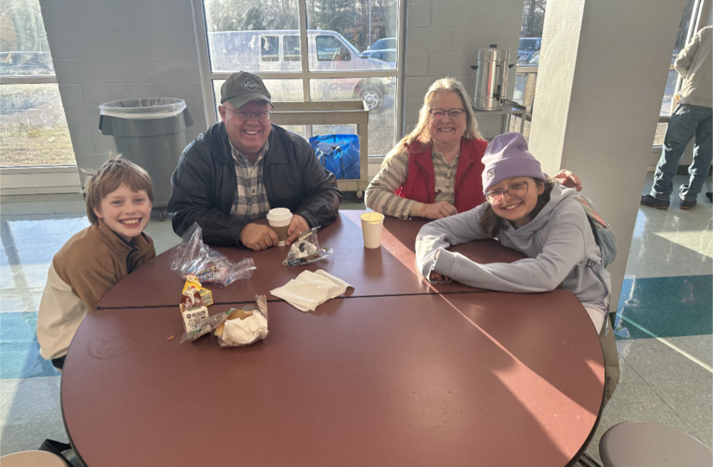 Students and their families sitting together at a table, smiling and enjoying muffins and coffee in the school cafeteria.