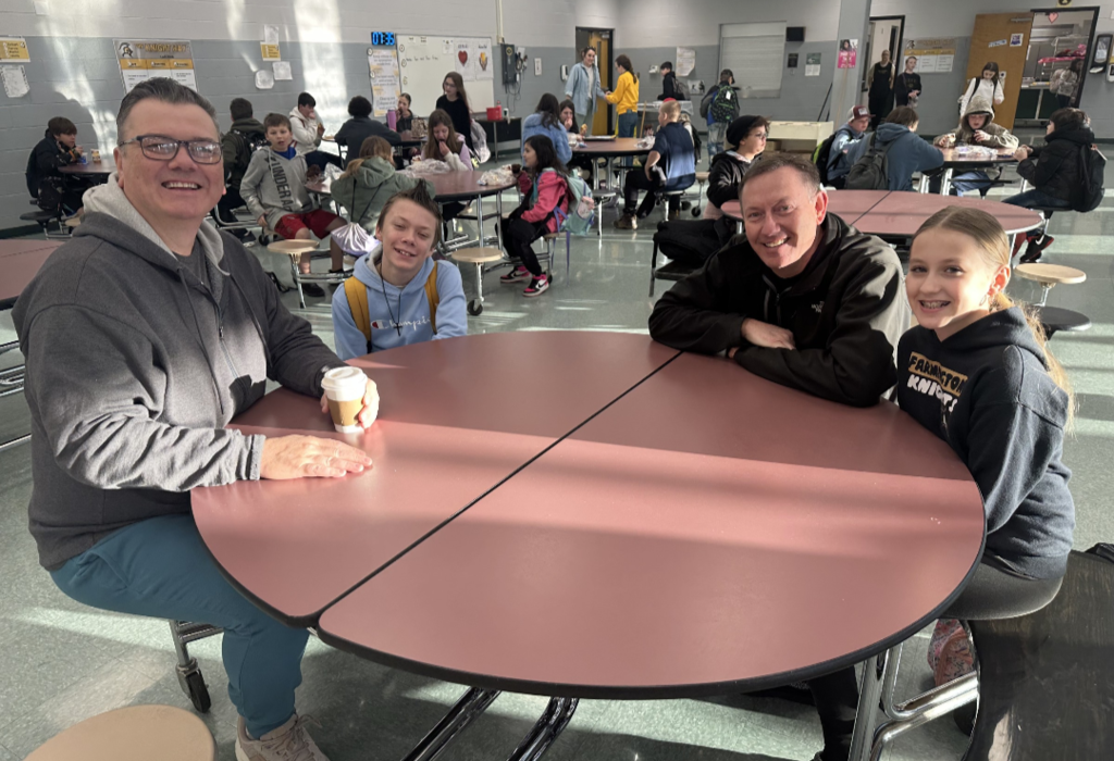 Students and their families sitting together at a table, smiling and enjoying muffins and coffee in the school cafeteria.