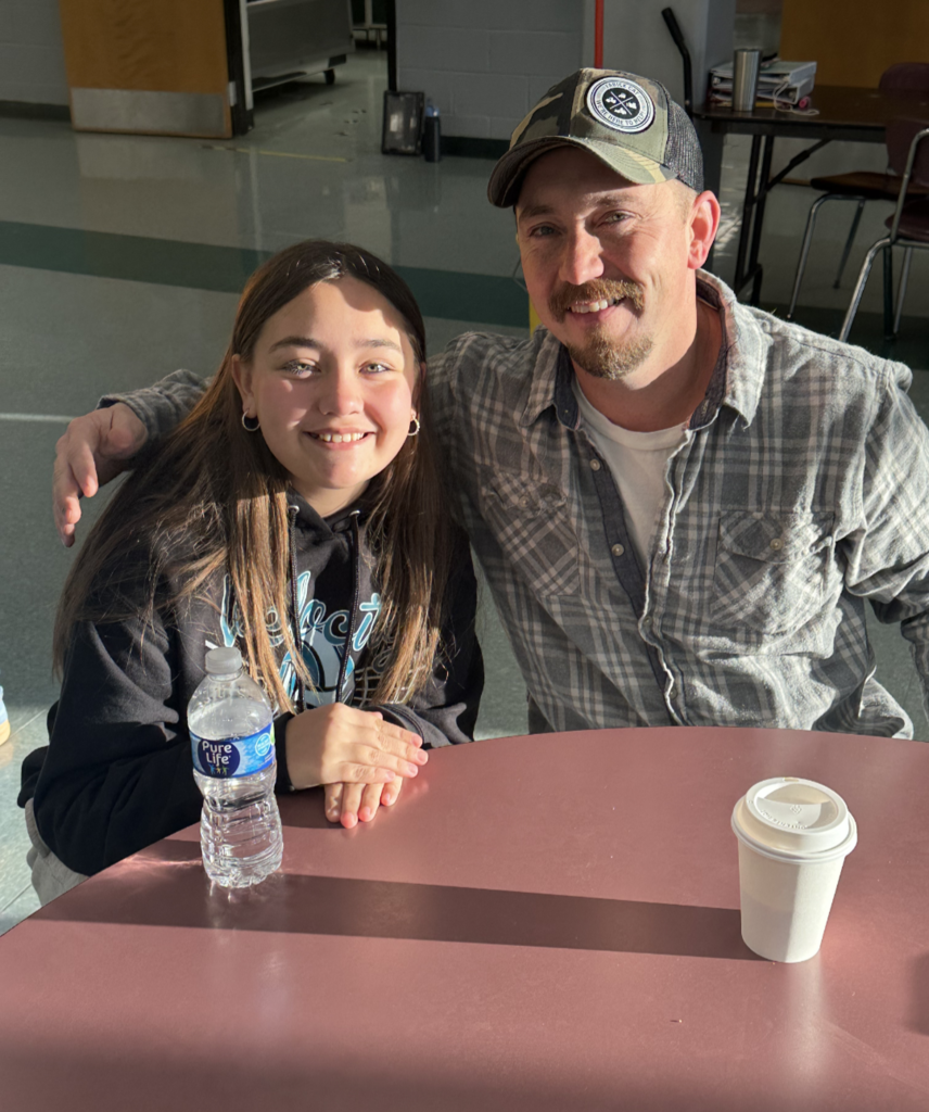 Students and their families sitting together at a table, smiling and enjoying muffins and coffee in the school cafeteria.