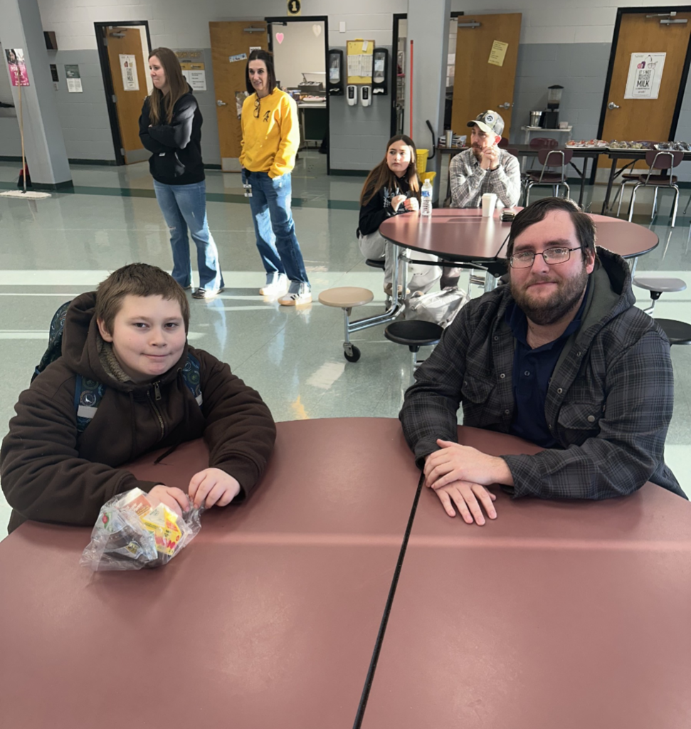 Students and their families sitting together at a table, smiling and enjoying muffins and coffee in the school cafeteria.