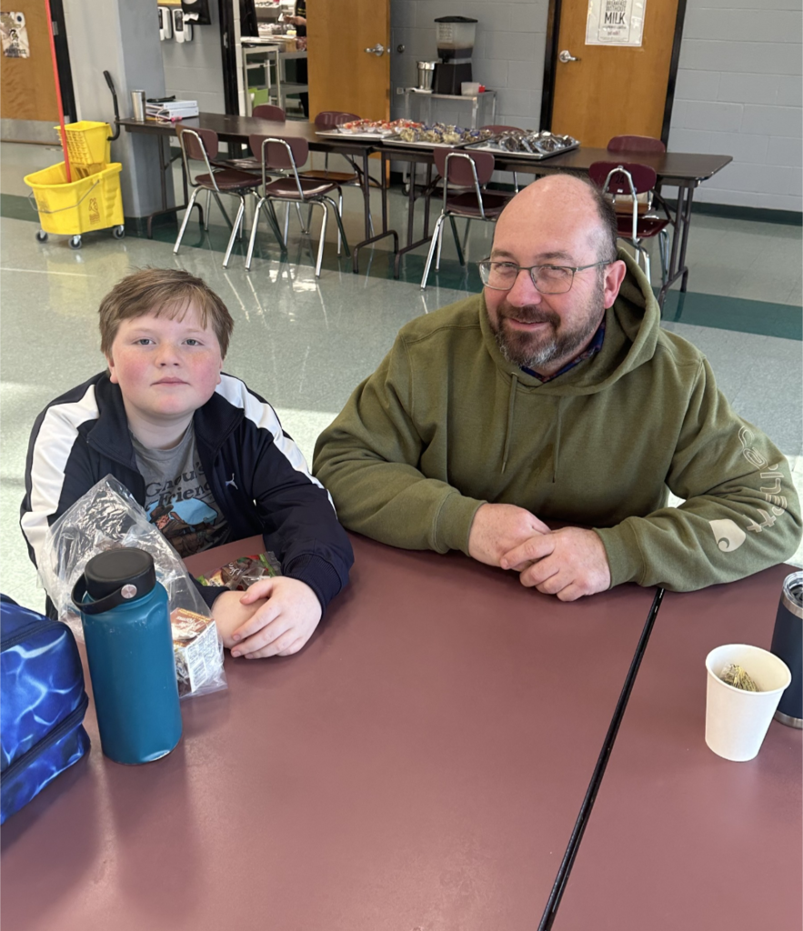 Students and their families sitting together at a table, smiling and enjoying muffins and coffee in the school cafeteria.