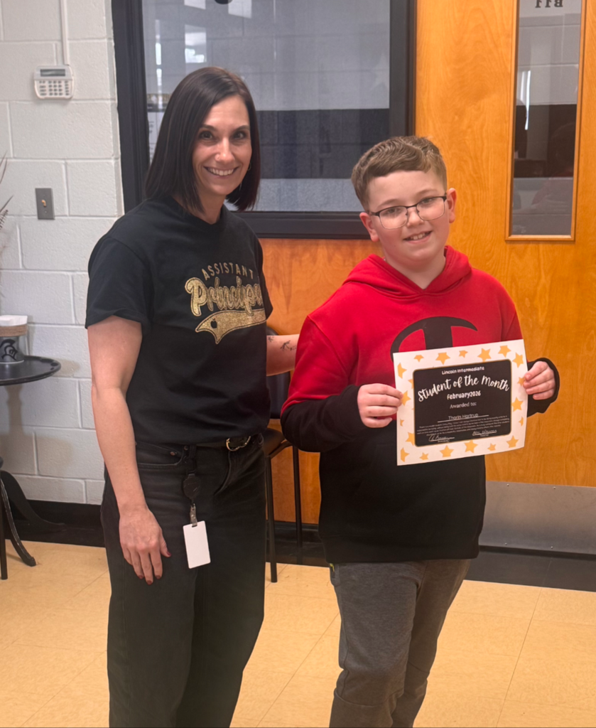 A smiling student and the assistant principal standing together indoors, holding a "Student of the Month" certificate and posing for a celebratory photo.