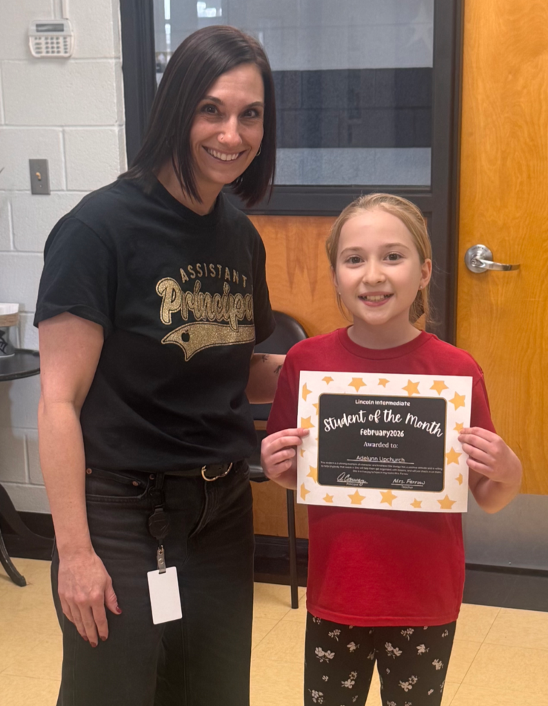 A smiling student and the assistant principal standing together indoors, holding a "Student of the Month" certificate and posing for a celebratory photo.