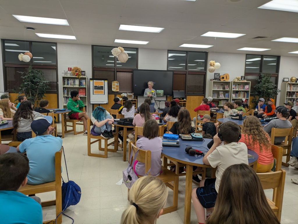 Local author Lisa Grider stands in a school library, smiling and gesturing as she speaks to an audience of students. She is holding a copy of her book, "Hobo’s Travels."  
