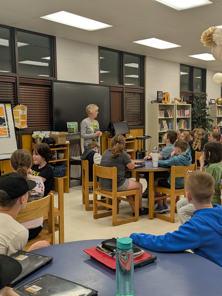 Local author Lisa Grider stands in a school library, smiling and gesturing as she speaks to an audience of students. She is holding a copy of her book, "Hobo’s Travels."