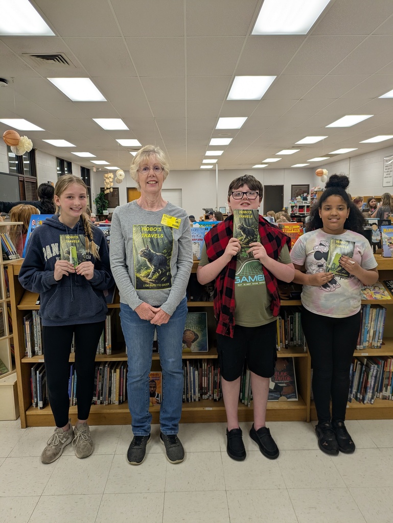 5th-grade students stand proudly in the school library, smiling and holding up signed copies of the book "Hobo’s Travels" alongside author Lisa Grider.