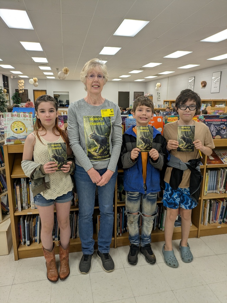 5th-grade students stand proudly in the school library, smiling and holding up signed copies of the book "Hobo’s Travels" alongside author Lisa Grider.