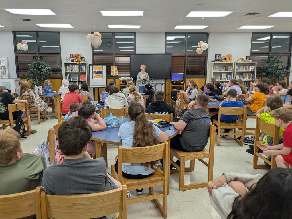 Local author Lisa Grider stands in a school library, smiling and gesturing as she speaks to an audience of students. She is holding a copy of her book, "Hobo’s Travels."