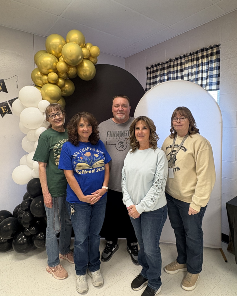 A group of five smiling school staff members pose together at a retirement celebration. They are standing in front of a festive backdrop featuring gold, white, and black balloon arches and circular display boards.