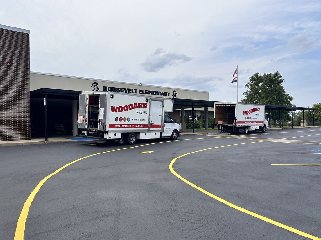 Two trucks with "WOODARD" on them, unloading in a school parking lot with two yellow lines.