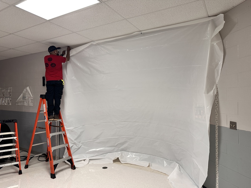 Person in red shirt and black pants on ladder, securing a white sheet to a wall in a room.