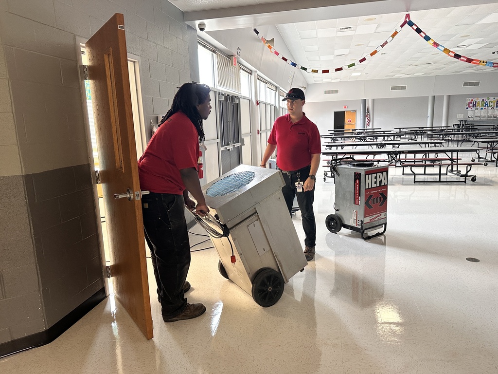 Two people in red shirts are pushing a large machine through a cafeteria with white floors.