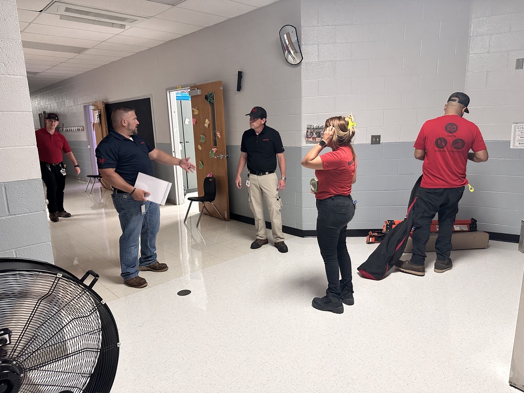 Five people in a hallway; a man holds a paper while gesturing, and three others look at him.