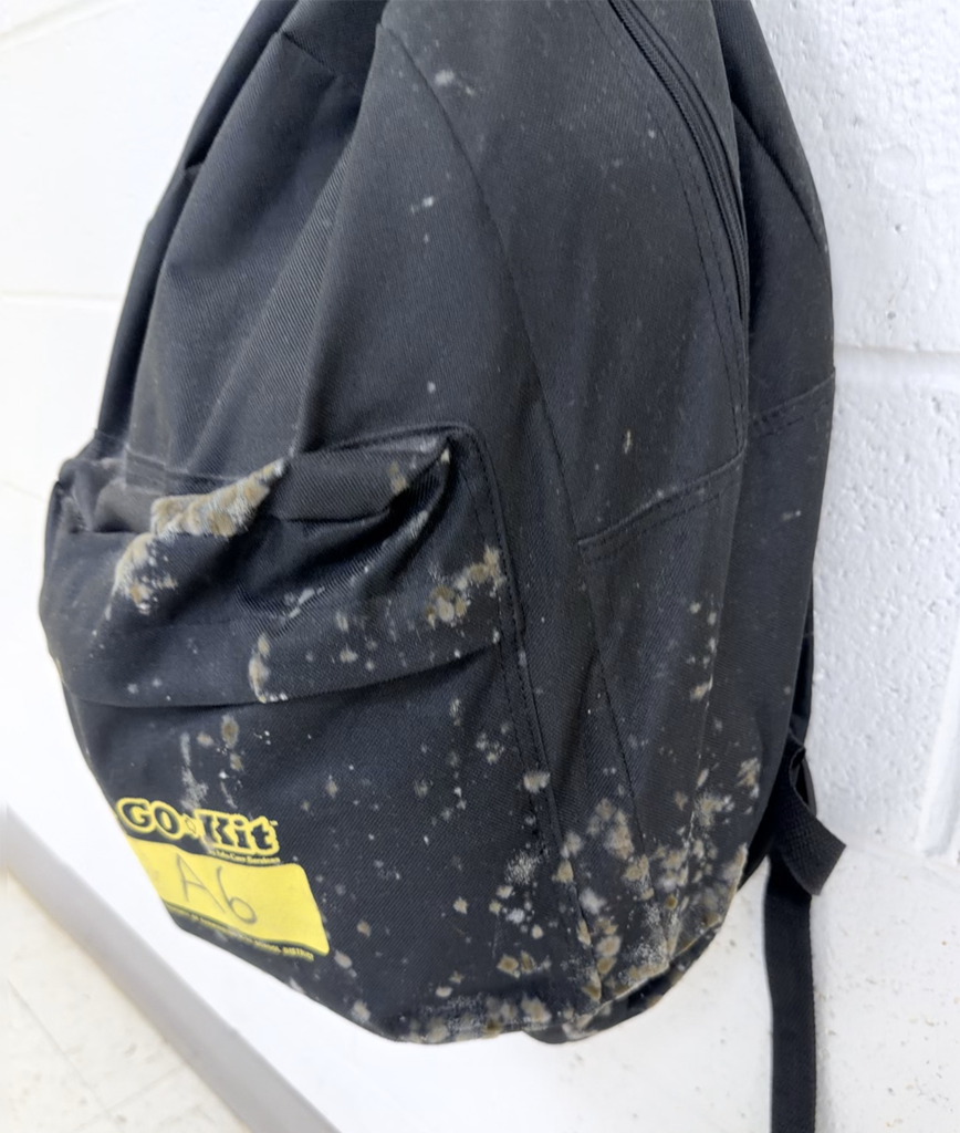 Black backpack with mud stains and a yellow label reading "Go-Rit 46" hanging on a white wall.