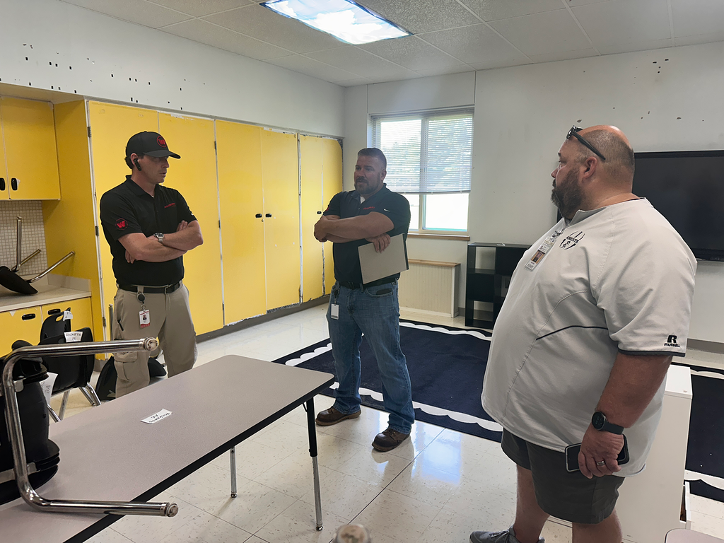 Three men in a room with yellow cabinets and a table, one holding a clipboard. One man wears glasses.