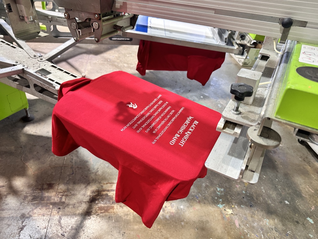 Red t-shirt being printed with text on a machine in a warehouse. Two other red shirts are also being printed.