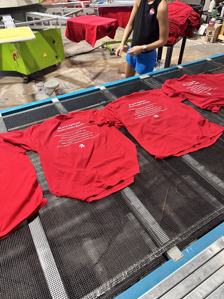 A woman stands near an industrial machine with four red T-shirts on it, in a warehouse.