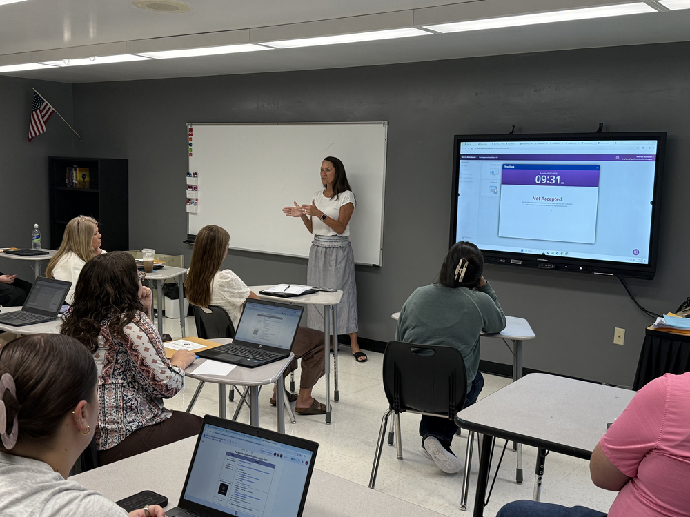 A classroom with students seated at desks and laptops. A woman stands at a whiteboard and speaks to the class.