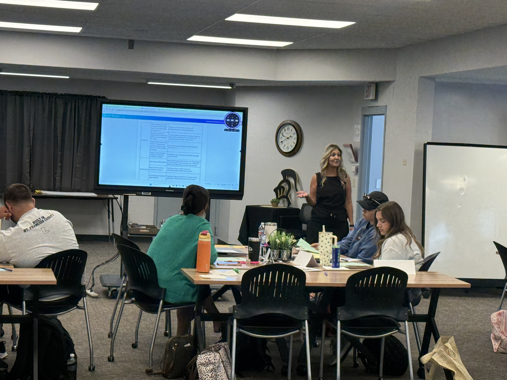 People sit in a classroom, listening to a woman at the front. A large screen displays behind her, and a clock adorns the wall.