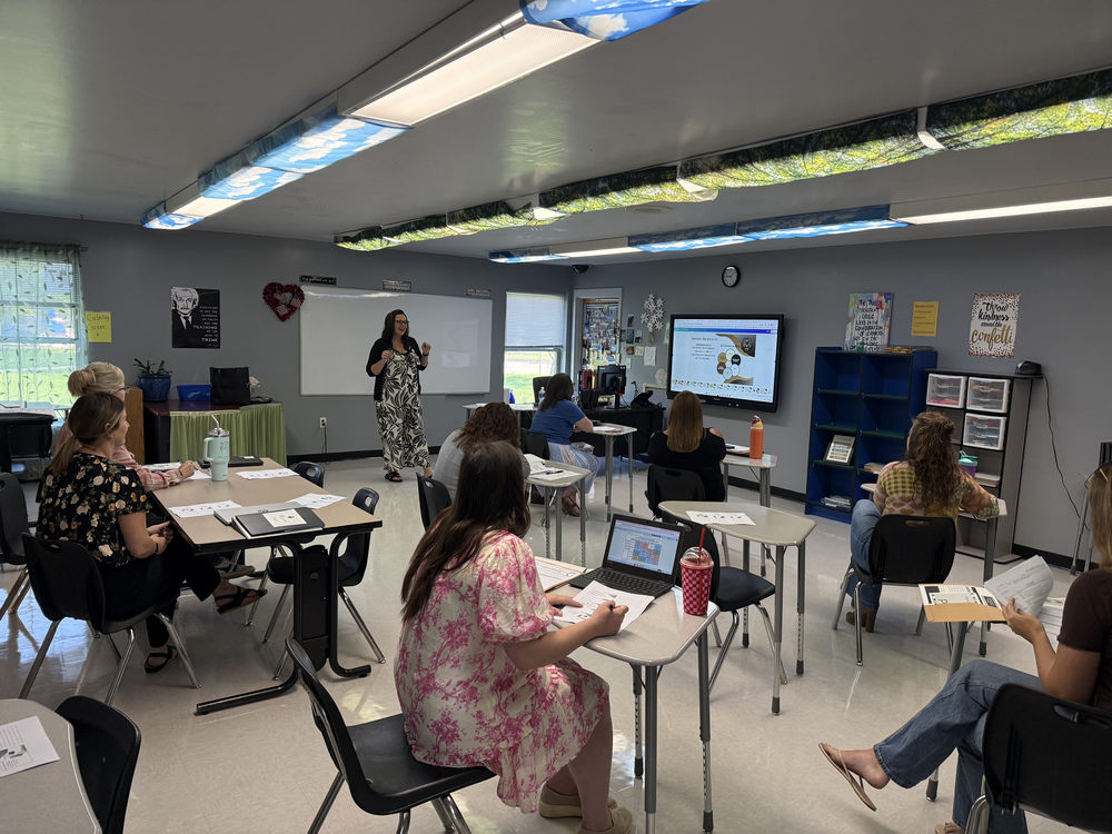 A classroom with a teacher at a whiteboard. Students in chairs around tables. White walls, bright ceiling lights.