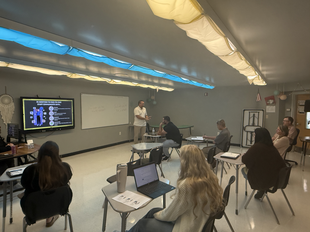 A classroom with multiple people seated around tables, listening to a man speaking at the front. A screen displays information.