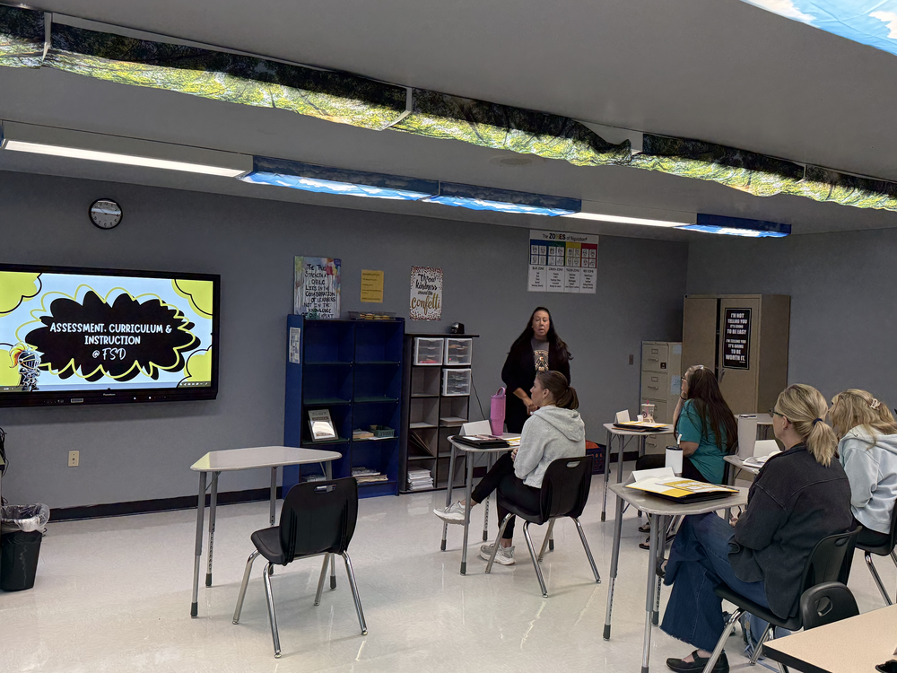 Classroom with fluorescent lighting, a large monitor on the wall, and five seated individuals. A woman stands in the center, facing the group.