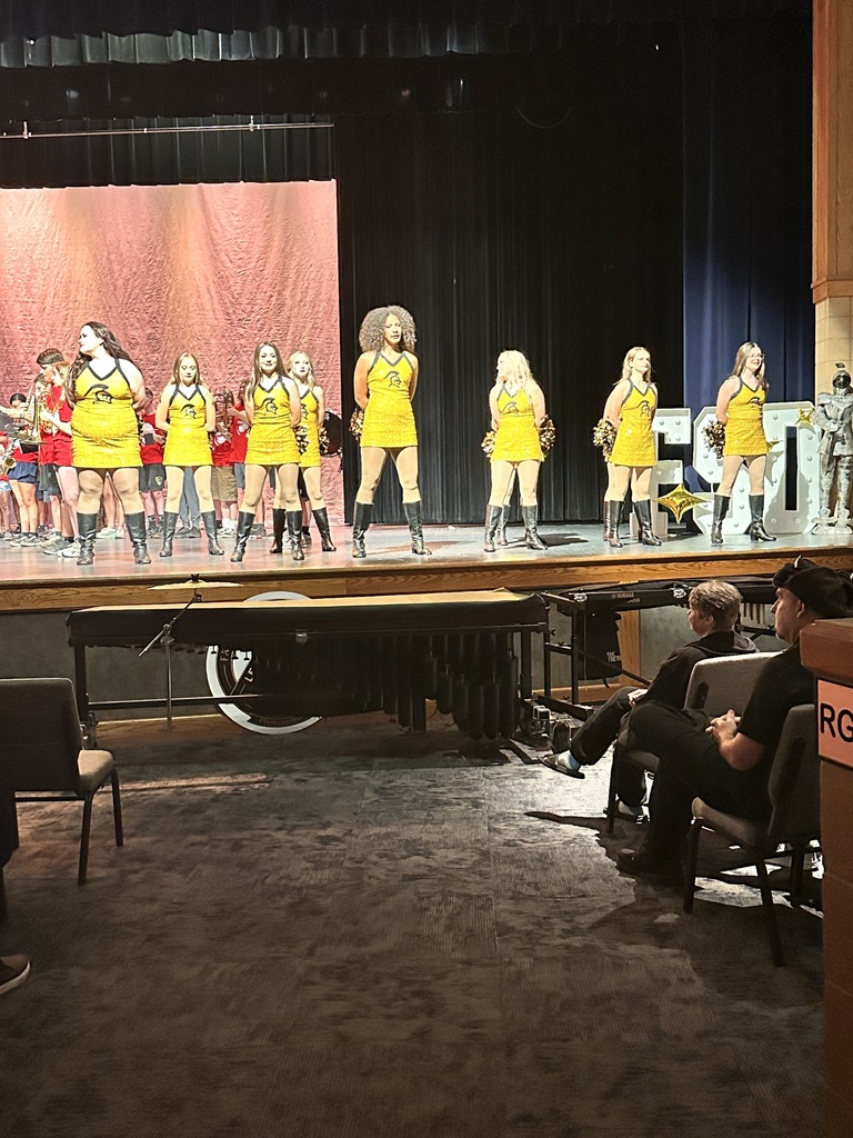 A group of cheerleaders in yellow outfits performing on a stage with a red backdrop.