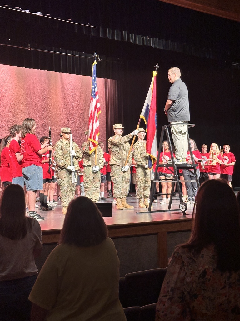 A man on stage with flags as soldiers stand behind him. People in red and white sit in the audience.