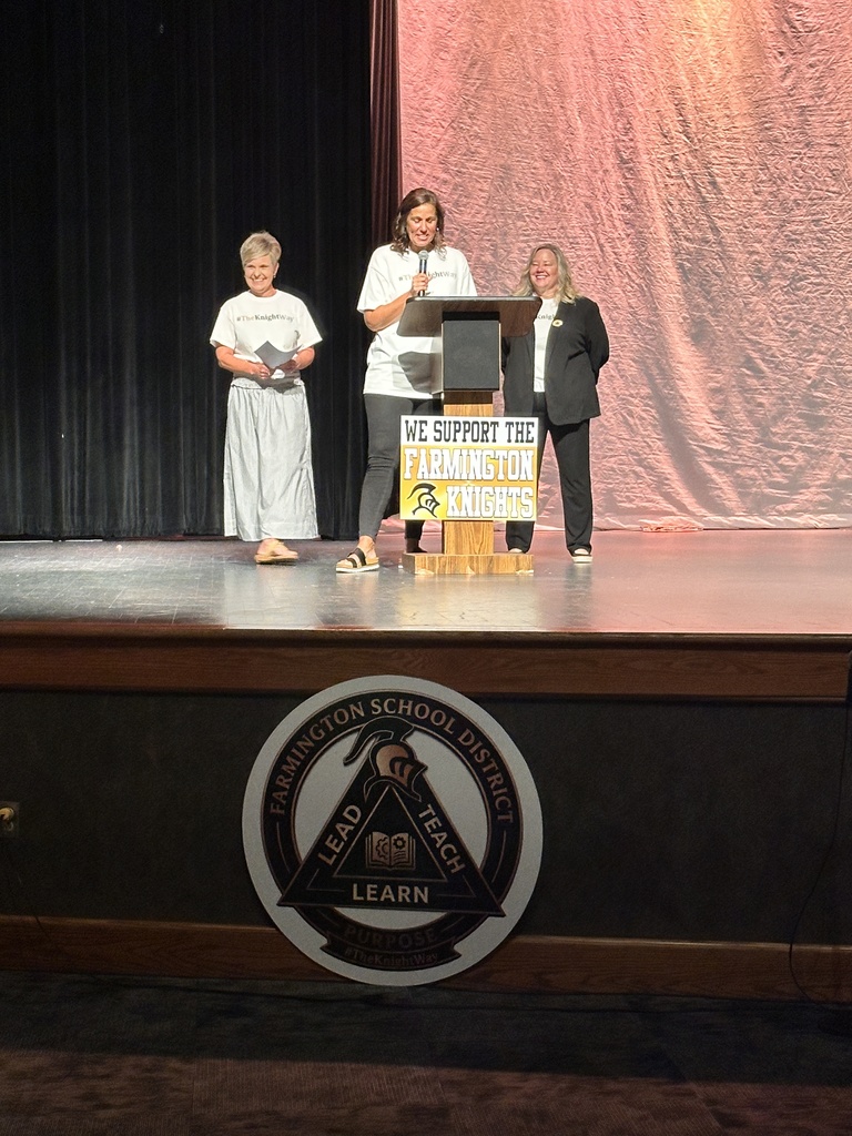 Three people on a stage, one speaking into a microphone. The stage has a logo and a backdrop.