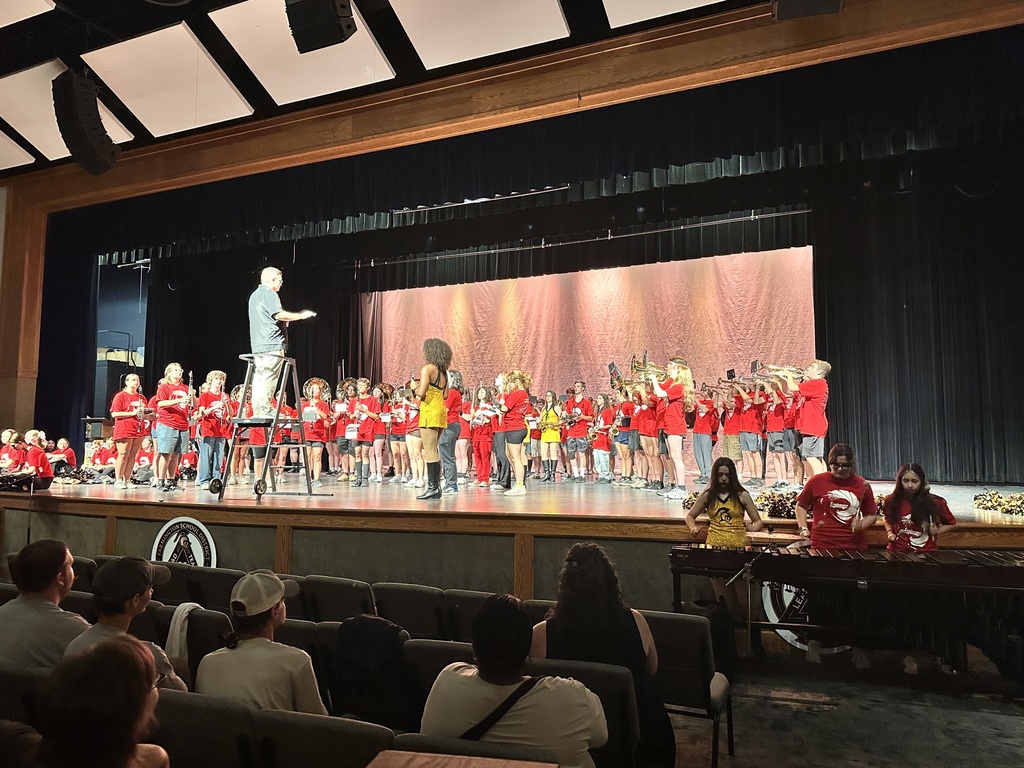 A group of people in red shirts performs on a stage. A person stands in front of them. Audience members sit in chairs in front of the stage.