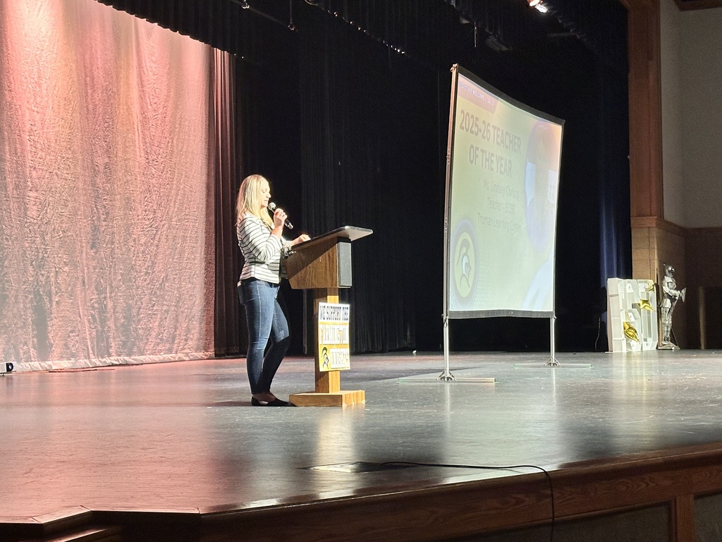 A woman with blonde hair stands at a podium with a microphone in a room with a projector screen.