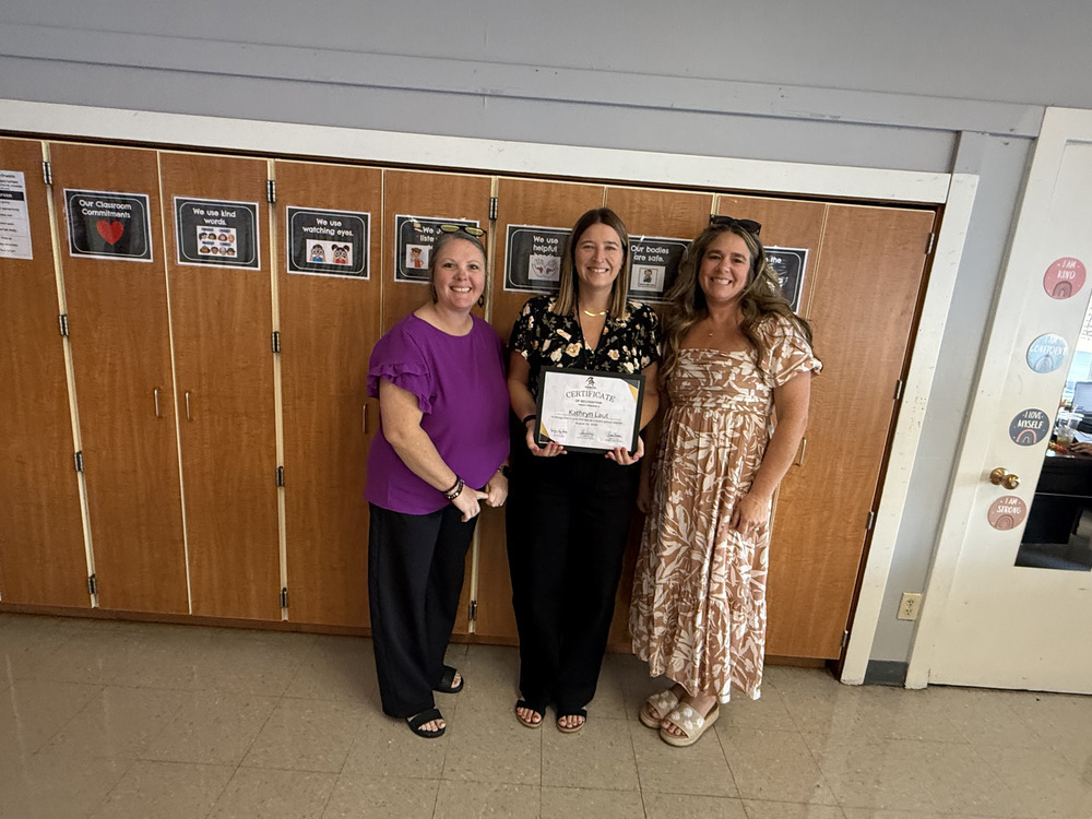 Three women stand together, one holding a certificate, in front of a wooden locker wall.